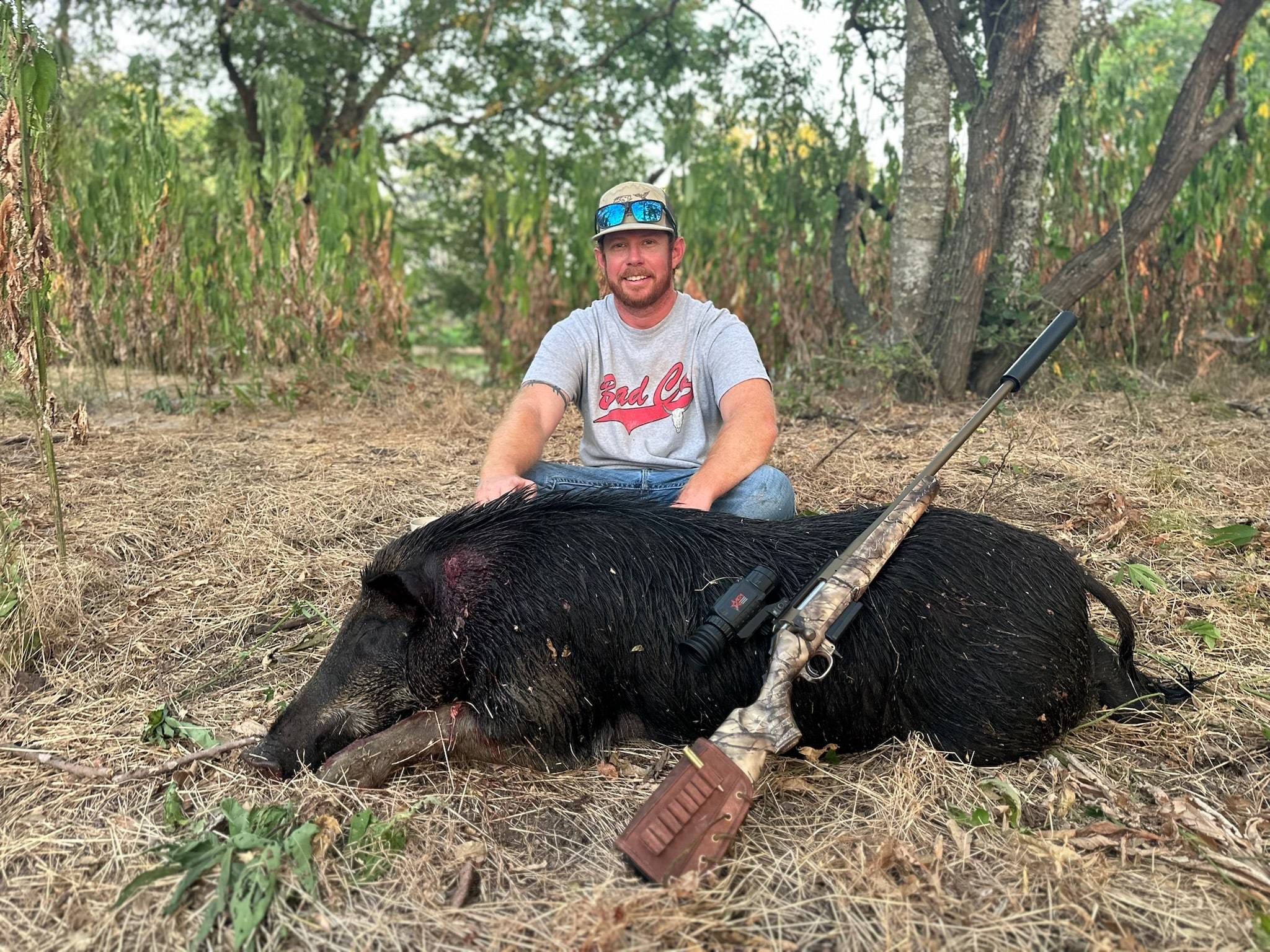 Hunter kneeling behind a large black feral boar with a suppressed bolt-action rifle and thermal scope laid across it