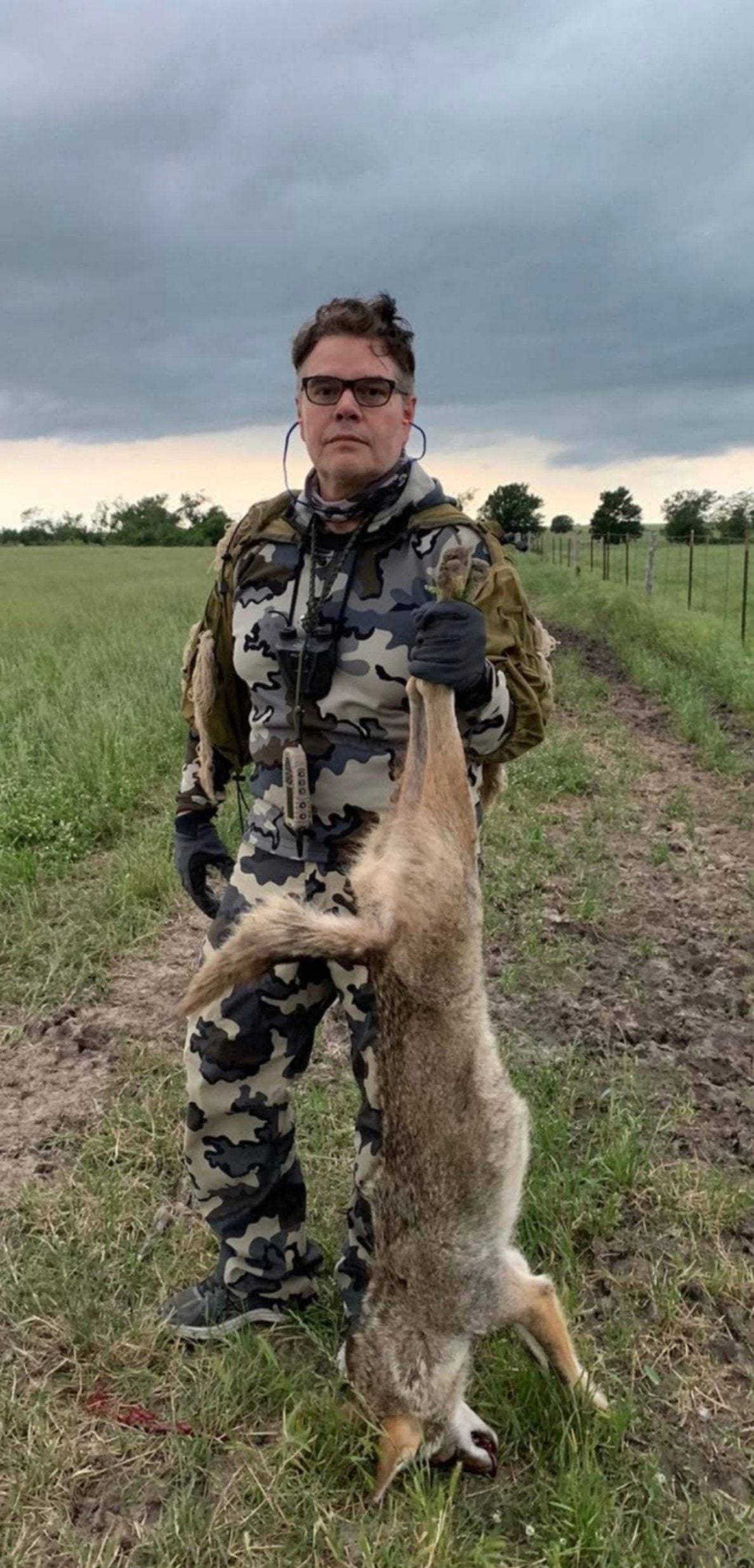 Hunter in gray camo with a predator call holding up a coyote on a pasture edge under storm clouds