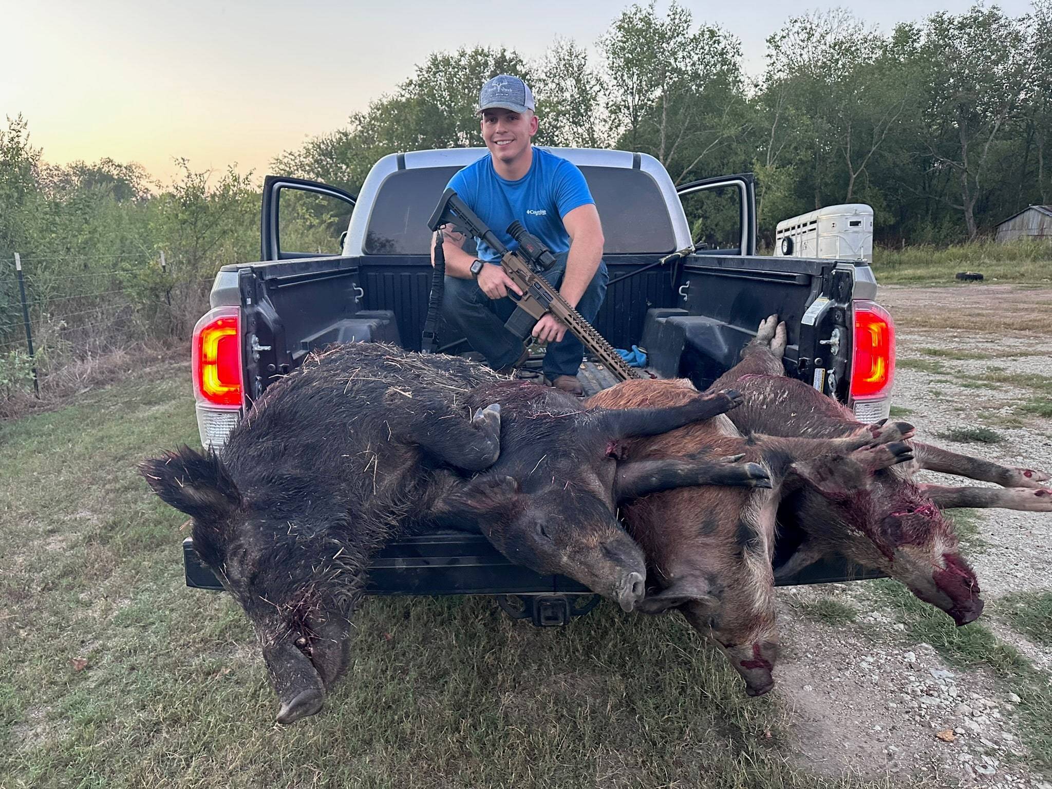 Hunter with a thermal-scoped AR rifle sitting in a pickup bed behind four feral hogs on the tailgate